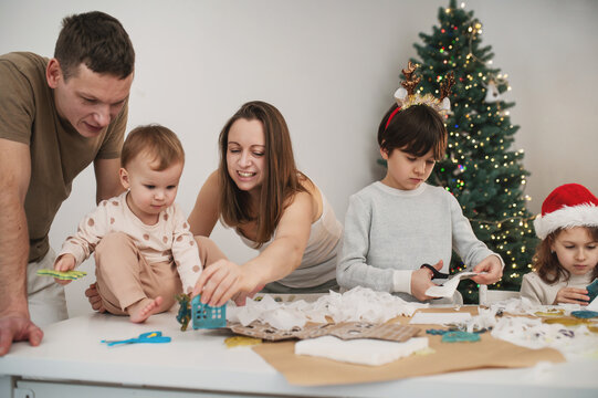 Parents and kids share creative holiday activity at home, cutting and making Christmas decorations together around table decorated with craft materials beside Christmas tree.