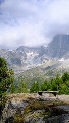 Fototapeta premium Banc en pierre face à un paysage alpin composé de conifères, de sommets rocheux et d’un glacier partiellement couvert par les nuages à Chamonix-Mont-Blanc, dans les Alpes françaises.