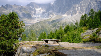 Naklejka premium Banc en pierre face à un paysage alpin composé de conifères, de sommets rocheux et d’un glacier partiellement couvert par les nuages à Chamonix-Mont-Blanc, dans les Alpes françaises.