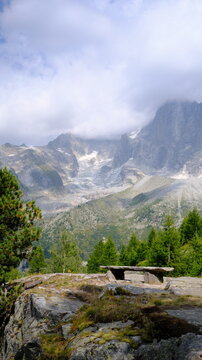 Banc en pierre face &agrave; un paysage alpin compos&eacute; de conif&egrave;res, de sommets rocheux et d&rsquo;un glacier partiellement couvert par les nuages &agrave; Chamonix-Mont-Blanc, dans les Alpes fran&ccedil;aises.