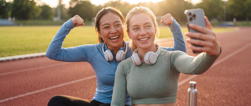 Fitness friends taking selfie on running track, cheerful diverse athletic women flexing muscles after sports workout at outdoor stadium in golden hour sunset, banner