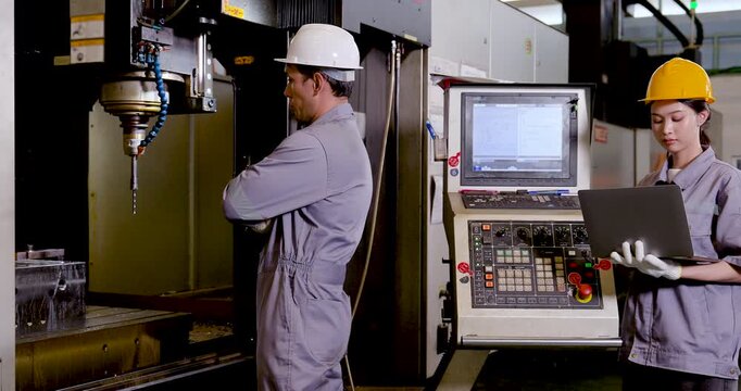 Asian man engineer studies CNC machine chamber while Asian woman technician holds laptop, tracks production data, and supports real time monitoring with safe teamwork in factory, slow motion