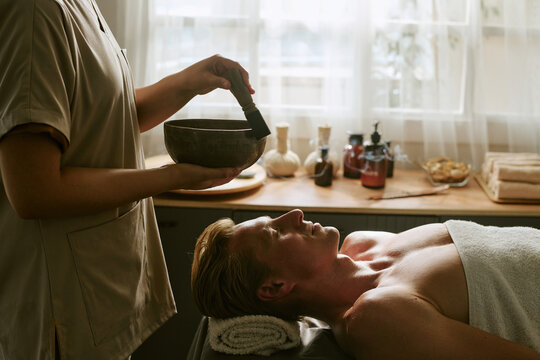 Masseuse using Tibetan singing bowl over Caucasian middle aged man lying on massage table during sound therapy session. Image suitable for wellness center and holistic spa marketing