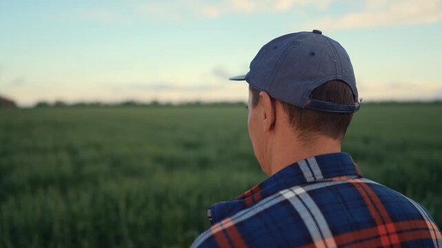 The man in a cap stands in a field. The man gazes at the green field under the sky, contemplating the landscape ahead.