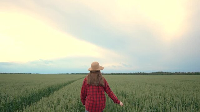 The woman walks through a green field, the woman wearing a red plaid shirt and a straw hat. The scene features a vast stretch of plants under a cloudy sky.