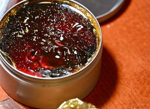 A commercial close-up photograph of an open round metal tin filled with dark, solid rosin flux used for electronics soldering and electrical work. The hard, amber-colored resin surface glistens under