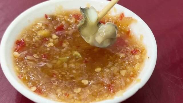 Close up of skewered spotted babylon snail moving over a bowl of spicy chili ginger fish sauce, Vietnamese street food