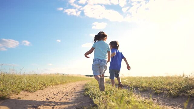 Two children, a girl and a boy, run together along a dirt path. The girl and the boy enjoy a sunny day in a grassy field.