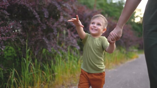 A woman and a boy walk together on a path surrounded by greenery and purple bushes. The boy smiles and holds the woman's hand as they enjoy the day.