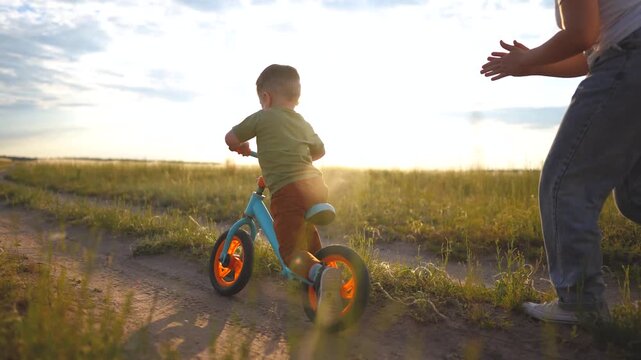A woman encouraging a boy riding a blue bike in a grassy field. The boy pedals his bike while the woman applauds his efforts.