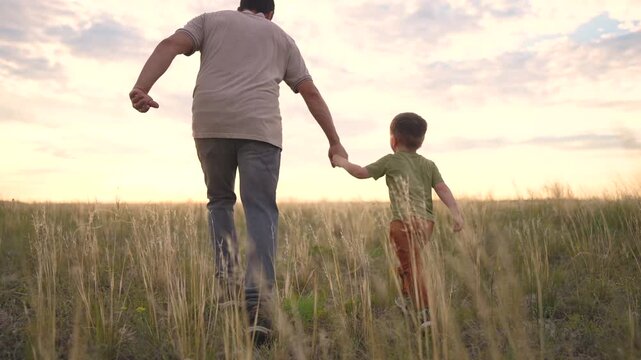 A man and a boy walk together in a field, holding hands. The man and the boy enjoy the view as they walk through the tall grass.