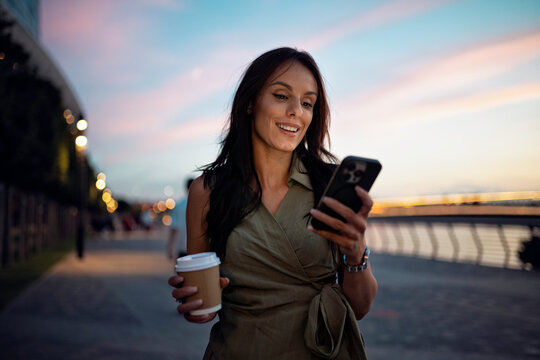 Woman smiling holding smartphone and coffee during sunset stroll