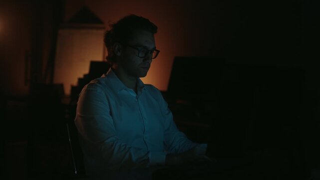 Focused man in glasses working overtime in dimly lit office, concentrating intensely on laptop screen with monitor cold light illuminating face against warm ambient glow, slow motion.
