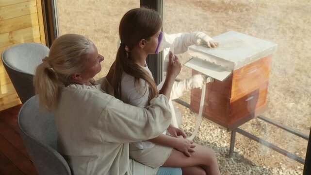 Young girl sits on her grandmother&rsquo;s lap and breathes beehive air through a mask while a beekeeper adjusts the airflow outside the apitherapy chamber.