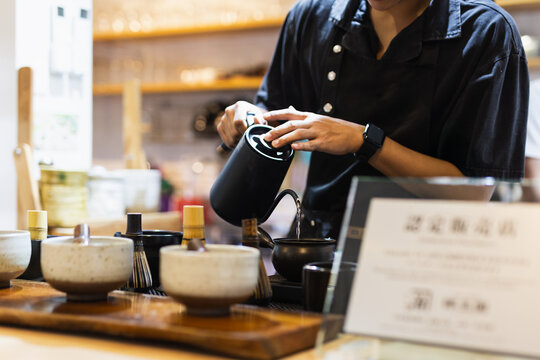 Barista pouring hot water from a black gooseneck kettle into a traditional teapot.