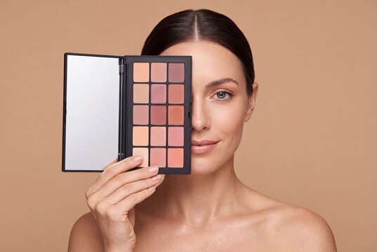 Woman holding a makeup palette covering half of her face against a neutral beige background