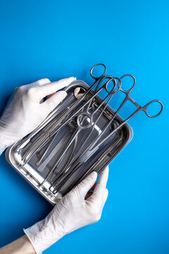 Hand in white glove is holding sterile surgical instruments and tools - scalpels, scissors and others - in a stainless tray, top view