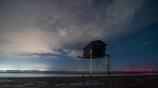A wooden stilt hut stands on a muddy beach under moving clouds and a starry sky at night in a coastal region of Vietnam.