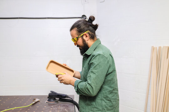 BEARDED CRAFTSMAN WEARING SAFETY GLASSES SANDING WOODEN TRAY IN WORKSHOP, STANDING AT WORKBENCH WITH POWER SANDER, WOOD MATERIALS, CLEAN WHITE BACKGROUND, HANDMADE FURNITURE PROCESS
