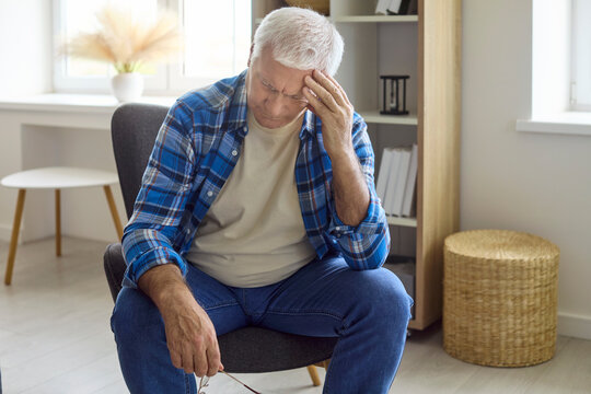 Senior man with headache feeling stress and pain. Sitting at home on a chair eyes down, showing fatigue and depression. Ideal for healthcare and anxiety ads. Clear concept of mental health.