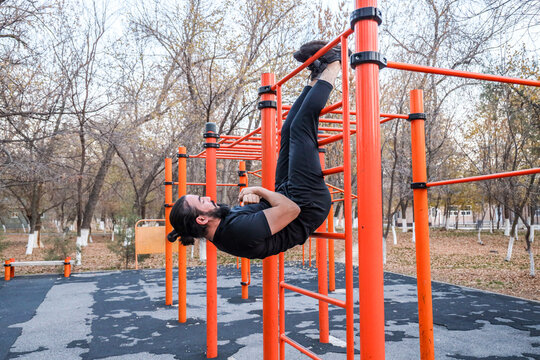 MAN PERFORMING HANGING LEG RAISE ON ORANGE CALISTHENICS BARS IN AUTUMN PARK, OUTDOOR FITNESS WORKOUT WITH STRONG CORE TRAINING, BODYWEIGHT EXERCISE, URBAN RECREATION, AND ACTIVE LIFESTYLE