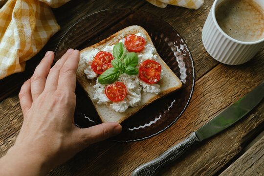 Woman having breakfast with a cup of coffee and sandwich. Hand holding toast topped with cream cheese, tomato and fresh basil leaves. Everyday routine, rustic style.