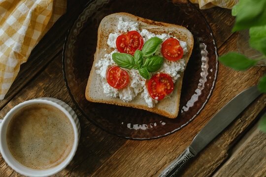 Breakfast with a cup of coffee and sandwich. Toast topped with cream cheese, tomato and fresh basil leaves on wooden table. Rustic style.