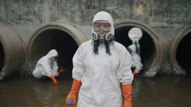 Environmental scientist in white protective gear looking confidently at the camera while monitoring water quality at a factory drainage outlet. Environmental research and pollution monitoring concept.