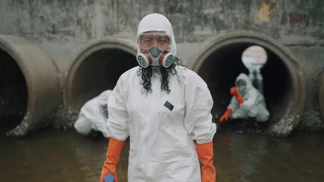 Environmental scientist in white protective gear looking confidently at the camera while monitoring water quality at a factory drainage outlet. Environmental research and pollution monitoring concept.