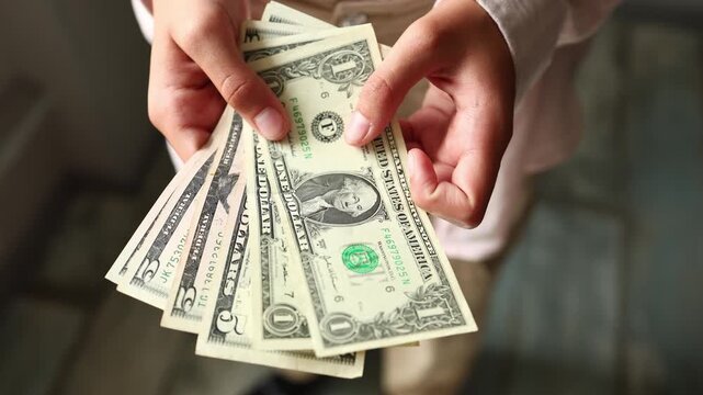 A girl is sitting in a room and counting small amounts of US dollars. She carefully sorts the cash in her hands. The light is soft as she focuses on the money.