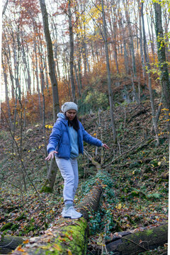 Adult woman carefully walks along a moss-covered fallen tree trunk in a deciduous autumn forest with arms extended for balance. Vertical outdoor scene illustrates adventure, agility, hiking, nature ex