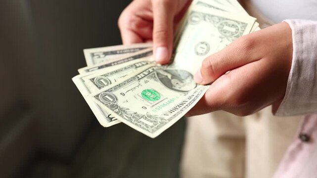A girl is sitting in a room and counting small amounts of US dollars. She carefully sorts the cash in her hands. The light is soft as she focuses on the money.