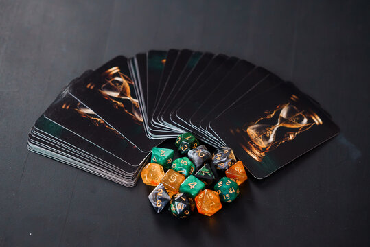 Pile of gaming cards with golden hourglass art and colorful polyhedral dice on a dark surface.