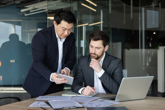 Two diverse businessmen are analyzing data and discussing strategy, collaborating in a modern office environment while utilizing digital technology and paper reports