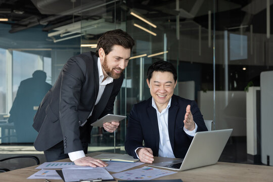 Two diverse business professionals are collaborating in a modern office, one man standing with a tablet and the other man sitting at a desk with a laptop, both discussing work and showing teamwork