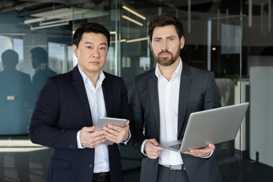 Two diverse businessmen in a modern office, one holding a tablet and the other a laptop, confidently posing to convey teamwork, partnership and digital business innovation