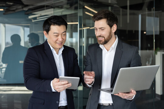 Two diverse businessmen standing in a modern office, analyzing data and discussing business strategies together using a digital tablet and laptop, fostering teamwork and professional development