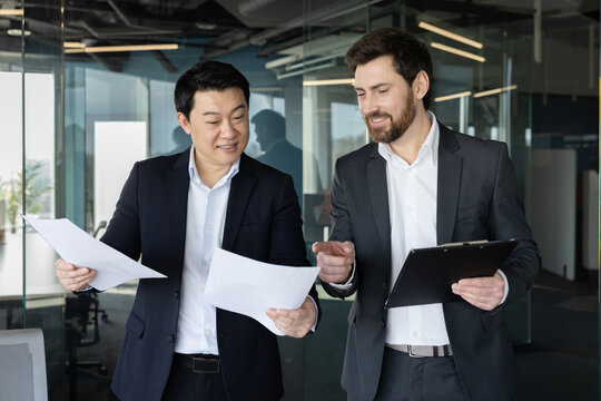 Two diverse businessmen in suits collaborating on documents, reviewing financial data, and discussing important reports during a corporate meeting in a contemporary workspace