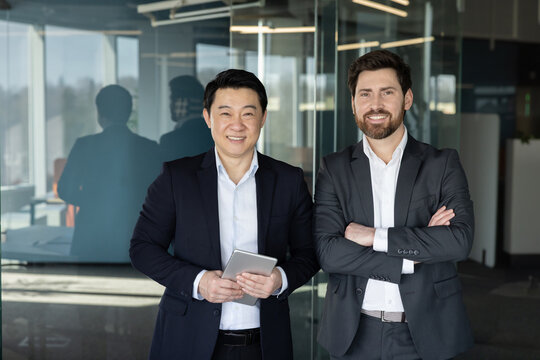 Two confident diverse businessmen, one holding a digital tablet, smiling and standing together in a contemporary office setting, representing successful teamwork and global partnership