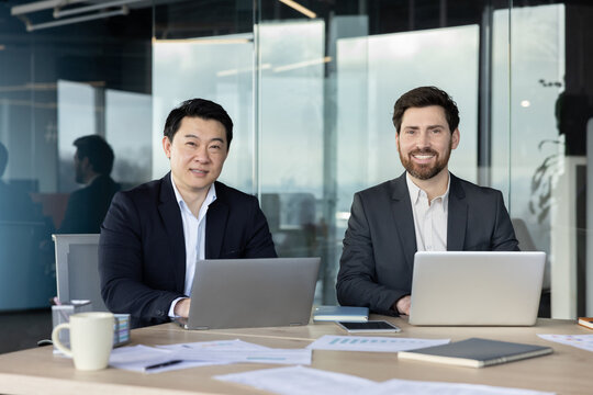 Two smiling multi-ethnic business partners sitting at a desk with laptops, collaborating on a project in a modern corporate office environment, representing professionalism and teamwork