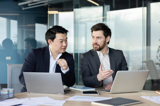 Two male business colleagues of different ethnicities are actively collaborating and brainstorming ideas during a professional discussion in a modern office. Using laptops and documents for their work