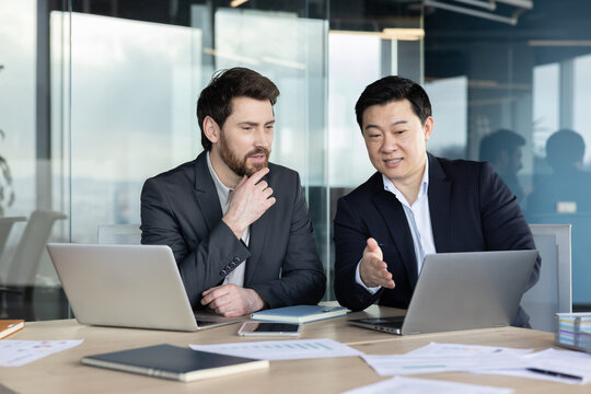 Two diverse business professionals are engaging in a focused discussion and collaborating on a laptop in a modern corporate office, planning strategy and sharing ideas