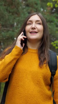 Cheerful young woman with a backpack walking through an autumn forest, smiling and talking on her smartphone while enjoying her hike and the beautiful natural scenery of the park