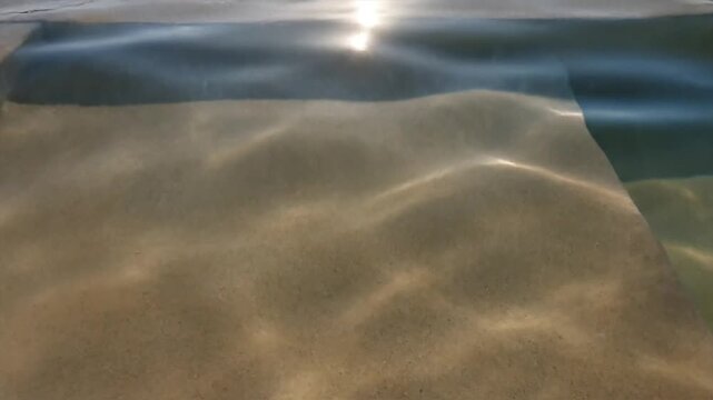 Sunlight caustic light patterns rippling across sandy pool floor through clear shallow water in slow motion