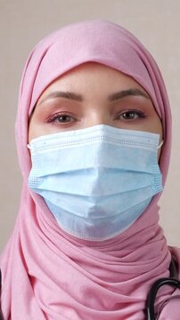 Close up portrait of a young muslim woman physician in a pink hijab and a protective face mask, wearing a stethoscope and looking directly at the camera with a confident expression