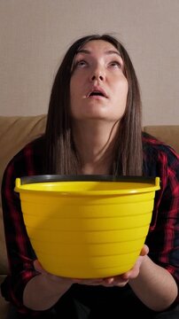 Worried woman sitting on her couch holding a yellow bucket, anxiously looking up as water drips from the ceiling into the container, showing the stress of a home emergency