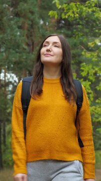Young serene woman in a yellow sweater with a backpack walking through a pine forest, enjoying the fresh air and beautiful nature during her hike on a beautiful sunny day