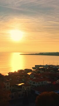 Sunset over coastal town and sea, Nesebar, Bulgaria. Aerial perspective of red roofs, marina entrance, calm water reflection and soft golden light at end of day. Vertical video.