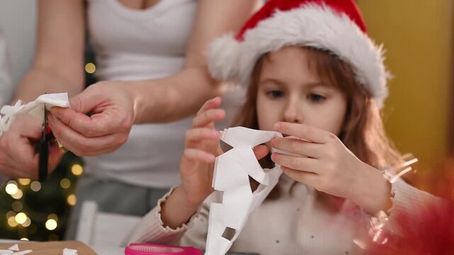 Mother helps 6 years girl makes paper snowflakes near a Christmas tree, celebrating Christmas with her family. New Year's preparations and decorations. Paper crafts. DIY