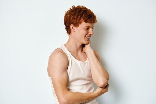 Young man with red hair wearing white tank top smiling and looking sideways. Casual portrait of male model with hand on neck and muscular arms against white background.